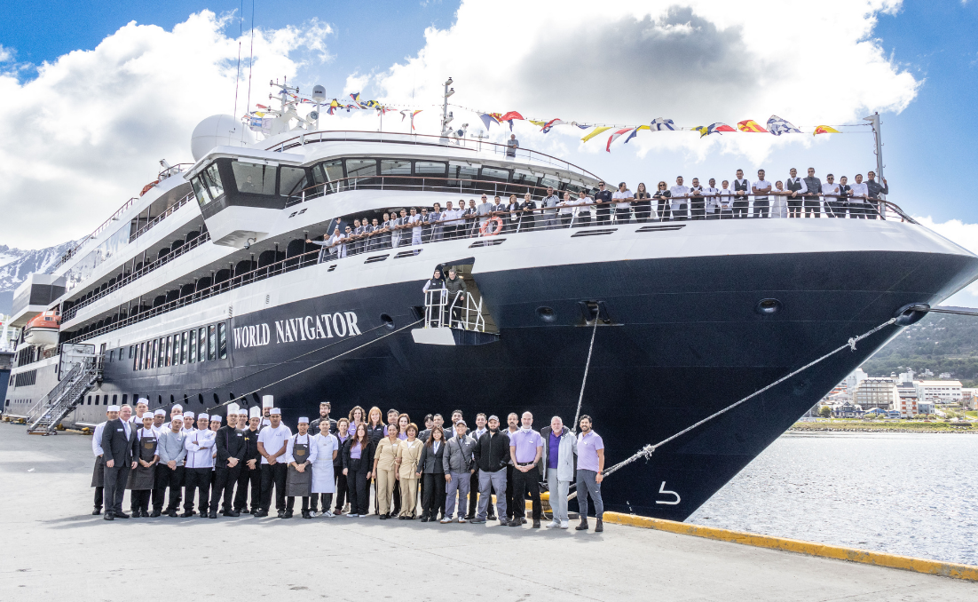 Tripulação do navio World Navigator reunida no cais e nos decks, com bandeiras náuticas no topo e o navio atracado ao fundo.