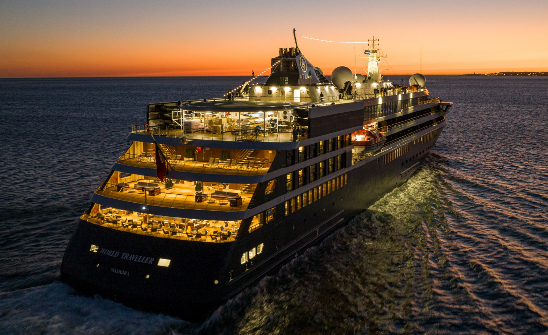 Navio de expedição World Traveller da Mystic Cruises a navegar ao pôr do sol, com os decks iluminados e rasto de água no oceano.
